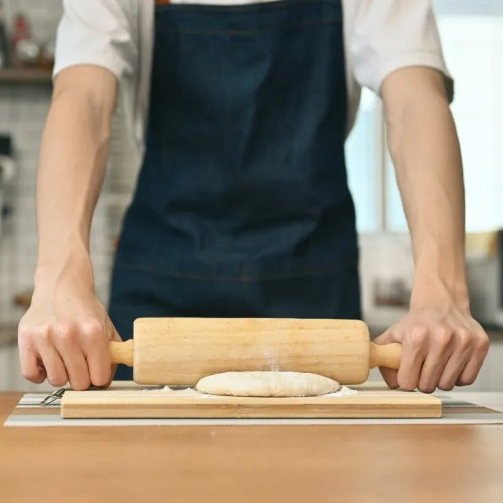 A man in an apron using a rolling pin to roll dough on a wooden board.