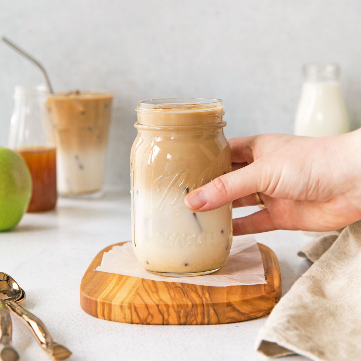 A hand holding a mason jar of iced coffee on a wooden coaster.