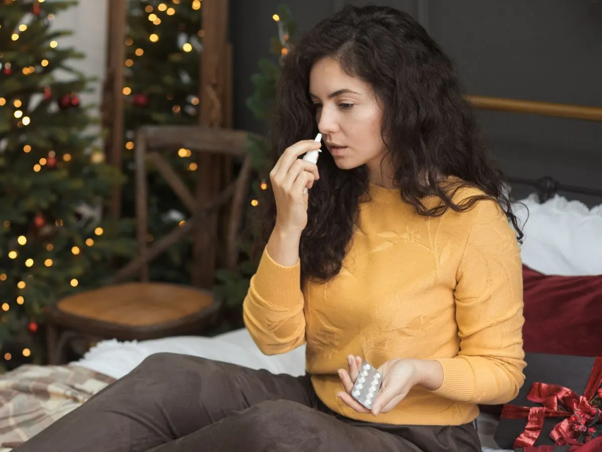 A woman is sitting on a bed in front of a Christmas tree using allergy medicine.