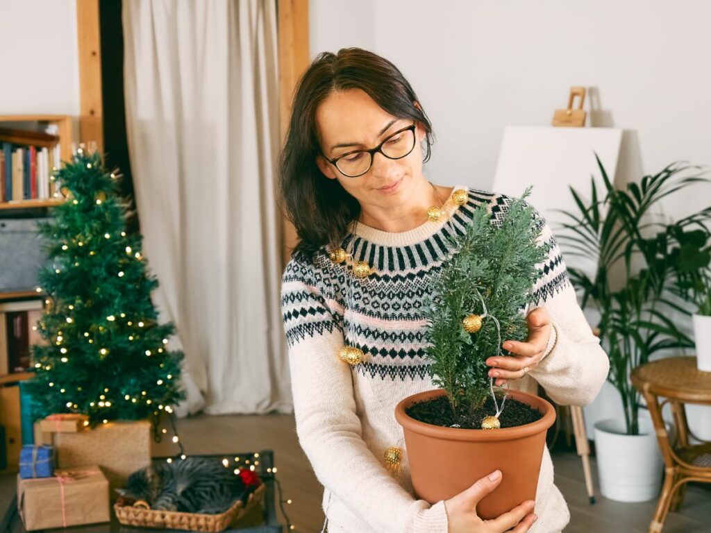 A woman holding a small Christmas tree in front of a larger Christmas tree.