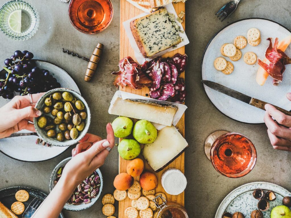 People's hands serving from a table with cheese, cold cuts, fruits, crackers and wine.