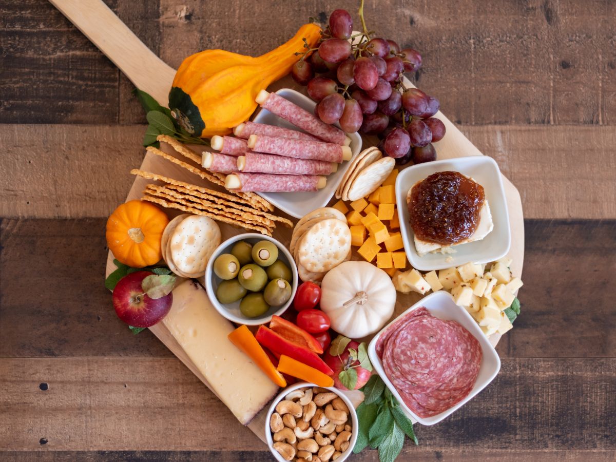 A wooden board with a variety of fruits, meats and cheeses on it.