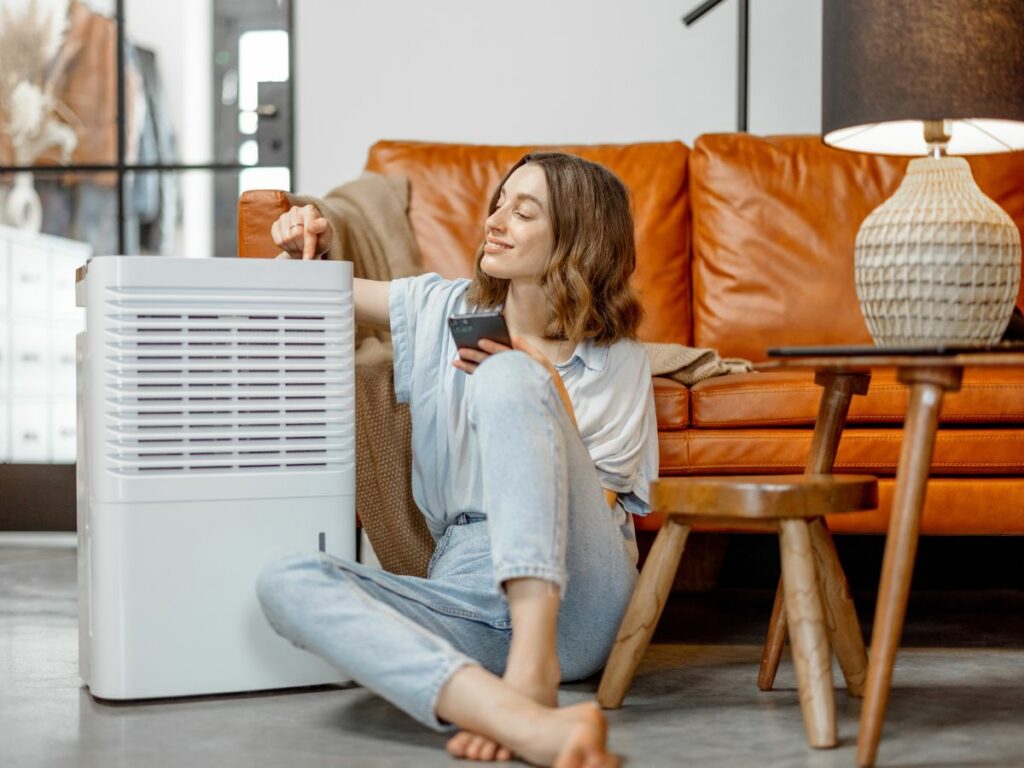 A woman touching a button while sitting on the floor next to a dehumidifier.