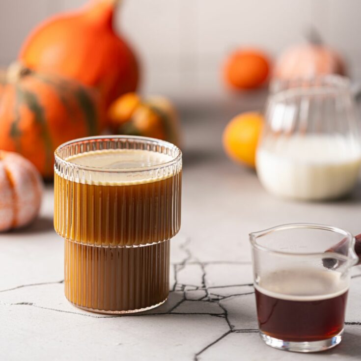 A cup of pumpkin latte next to various pumpkins and a shot of espresso.