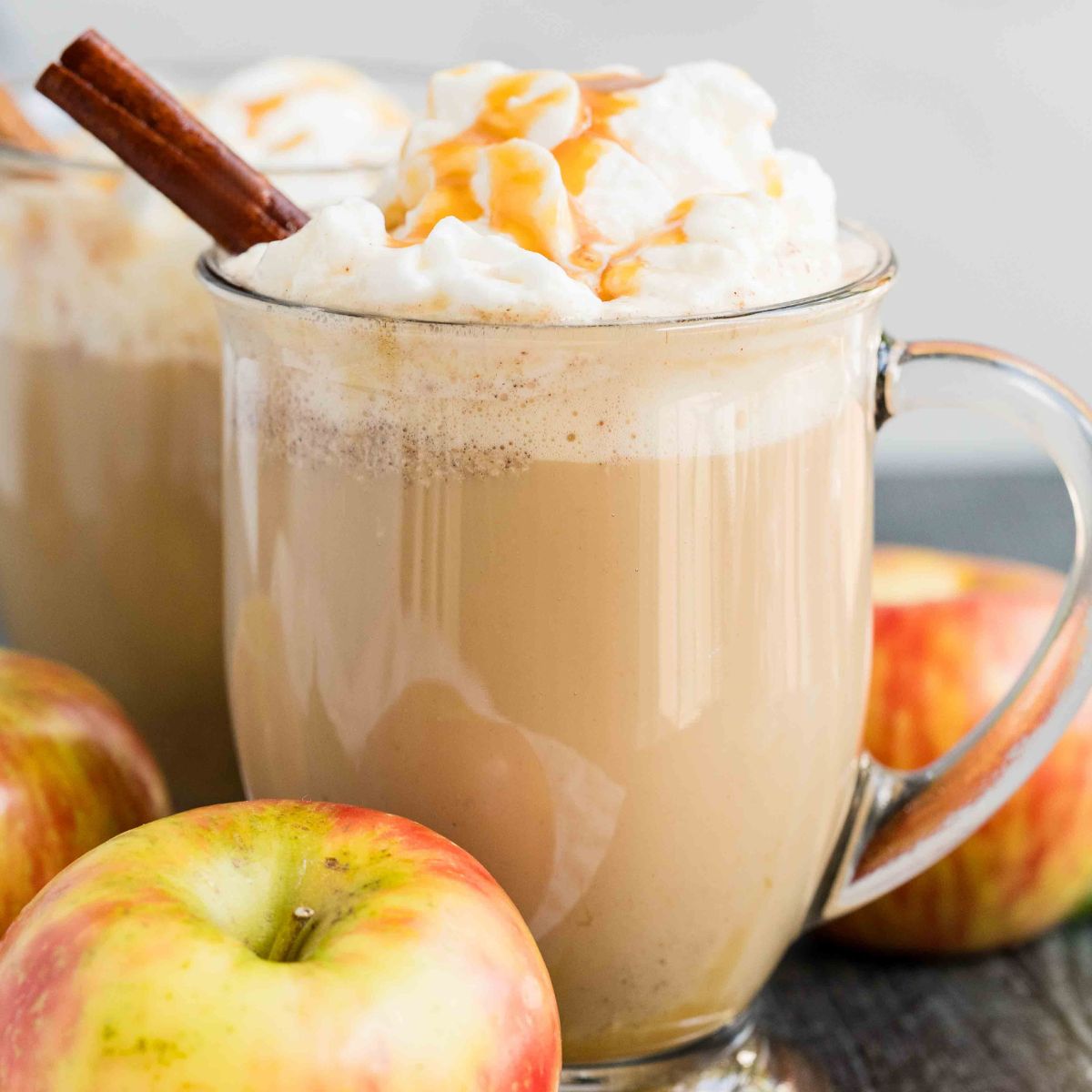 Two mugs of coffee with whipped cream next to apples.