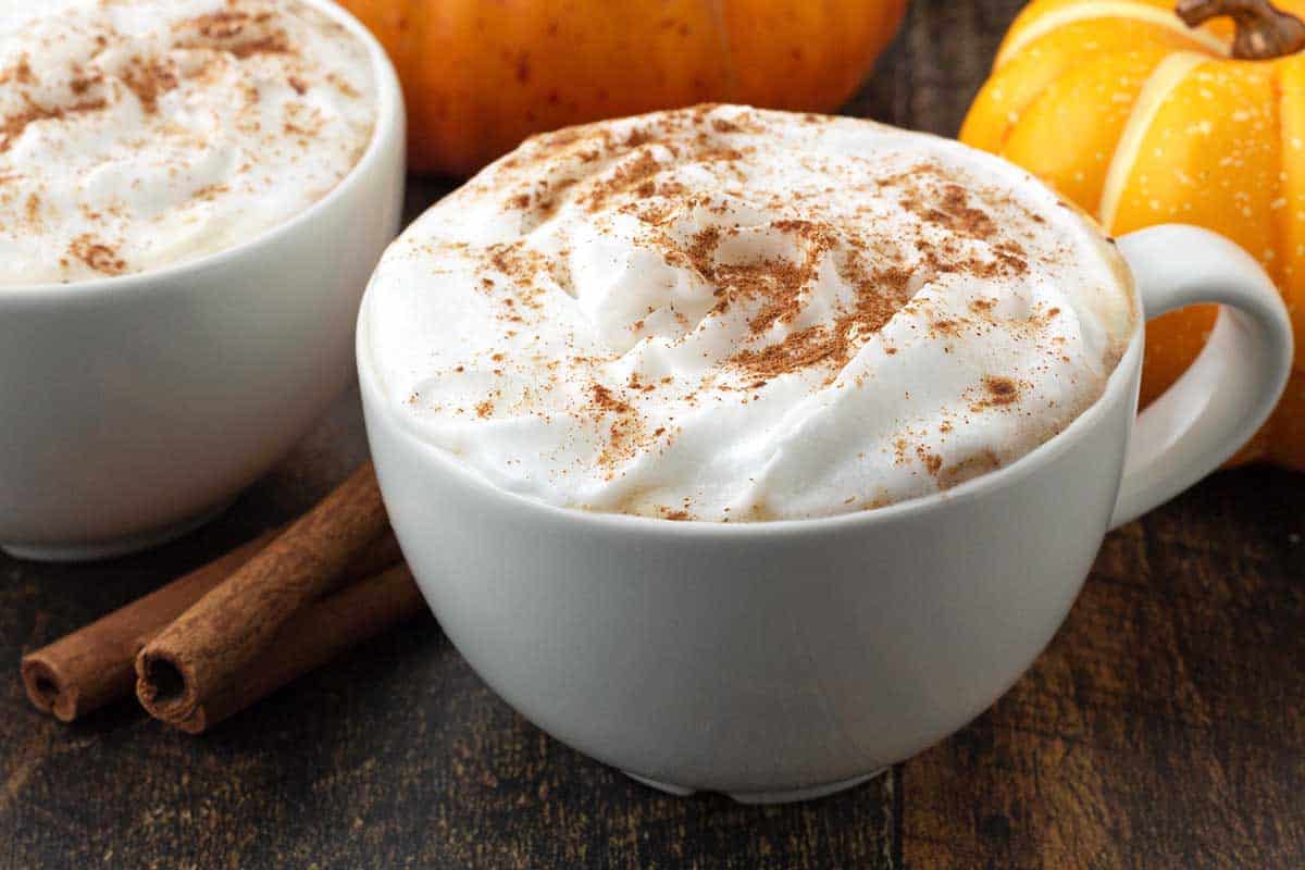 Two mugs of latte next to cinnamon sticks and pumpkins.