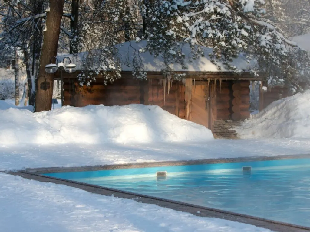 A wooden poolhouse in the snow with a warm pool in front.