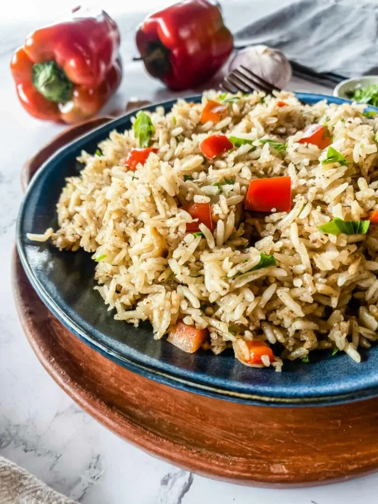 A blue plate with rice and peppers on it. A head of garlic and two red peppers on the background.