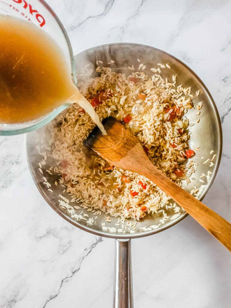 Chicken broth being poured into a skillet filled with rice.