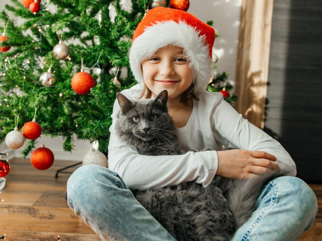 A girl in a santa hat sitting with a cat in front of a christmas tree.