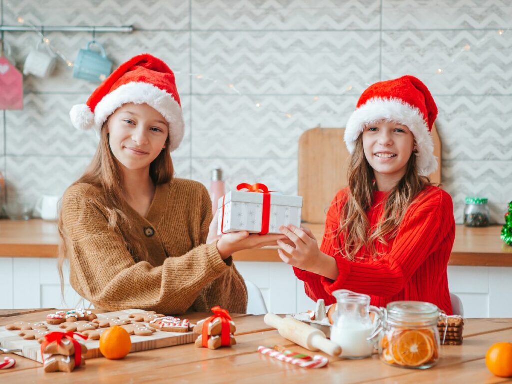 Two girls in Christmas hats passing a gift in the kitchen.