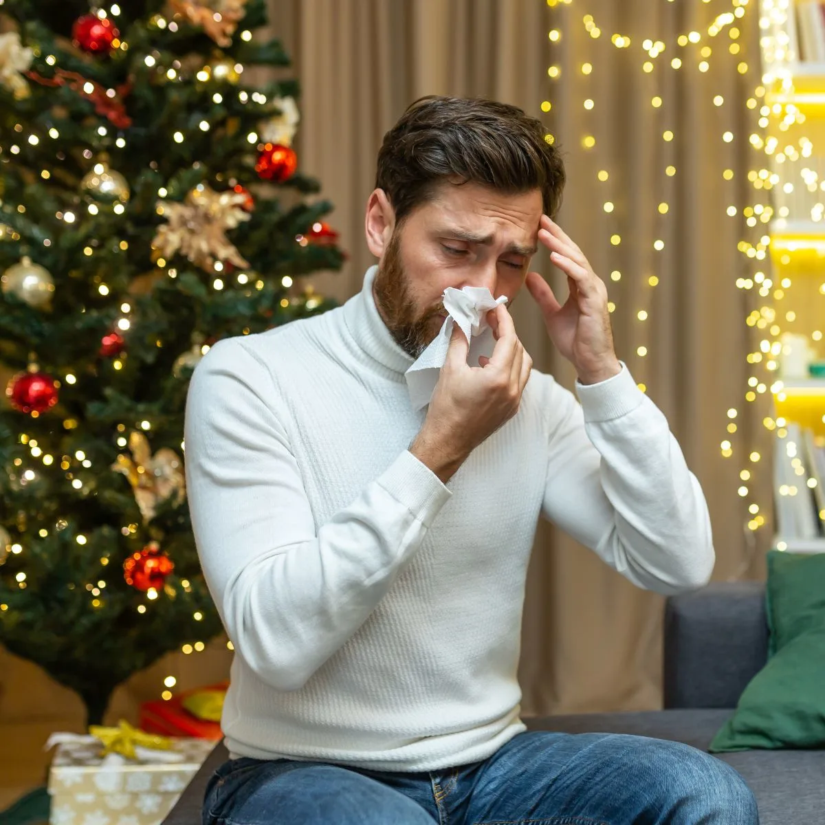 A man blowing his nose in front of a christmas tree.