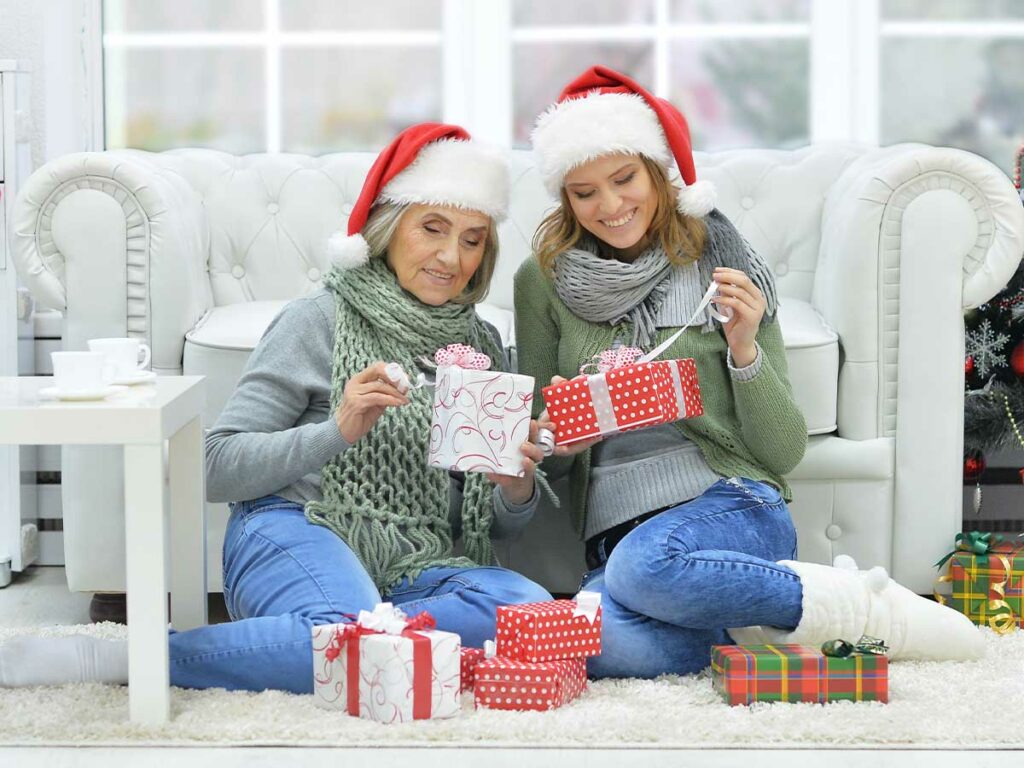 Two older women in santa hats sitting on the floor, exchanging Christmas gifts.