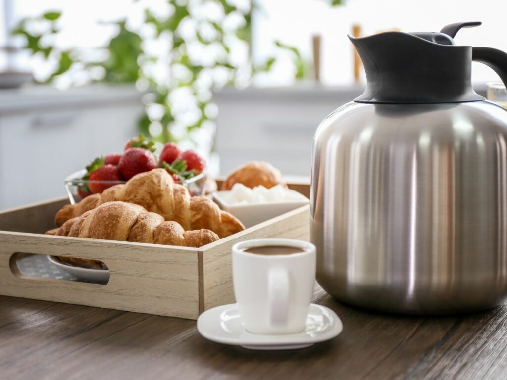A stainless steel coffee carafe on a tray with croissants and a cup of coffee.