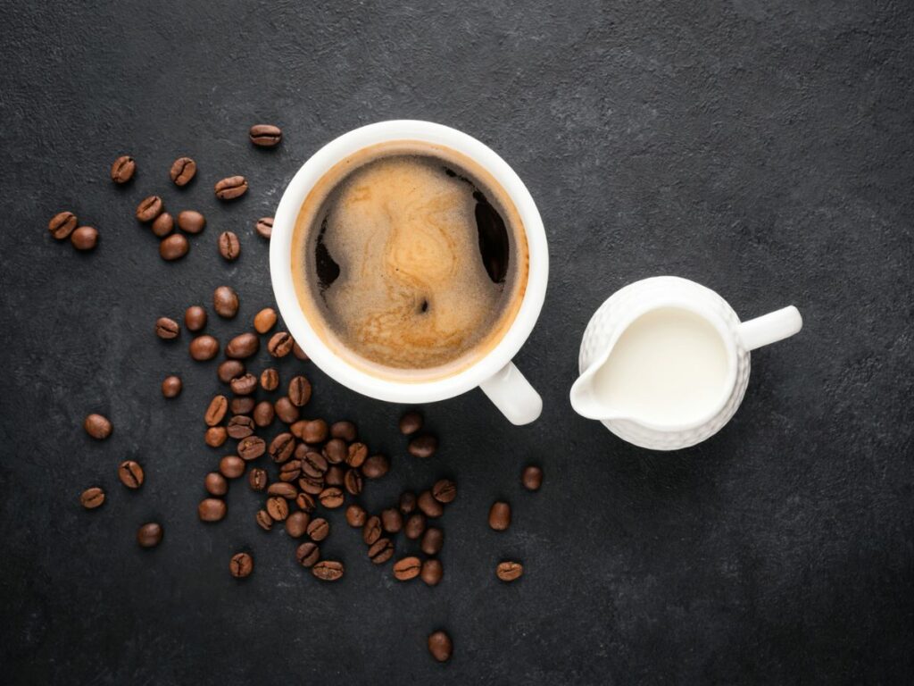 A cup of coffee with eggnog creamer on the side and coffee beans on a dark background.
