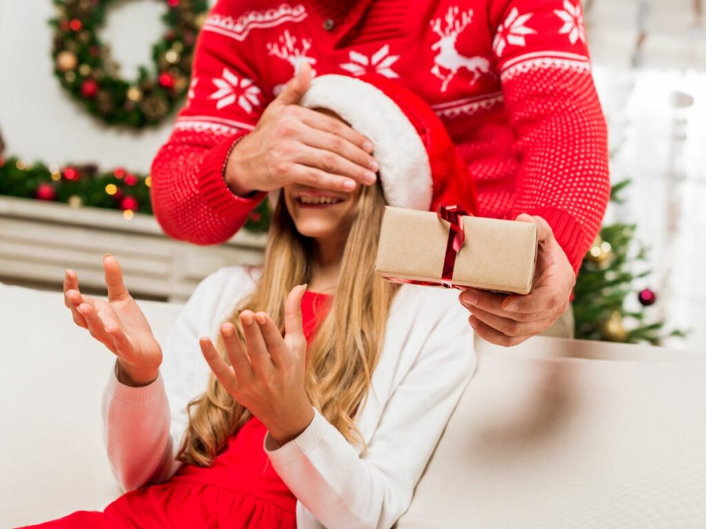 A man is holding a gift box in front of a teen girl in a santa claus hat.