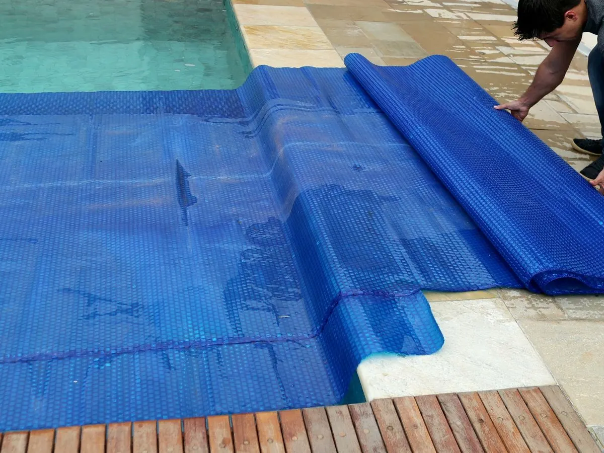 A man laying down a blue pool cover on a wooden deck.