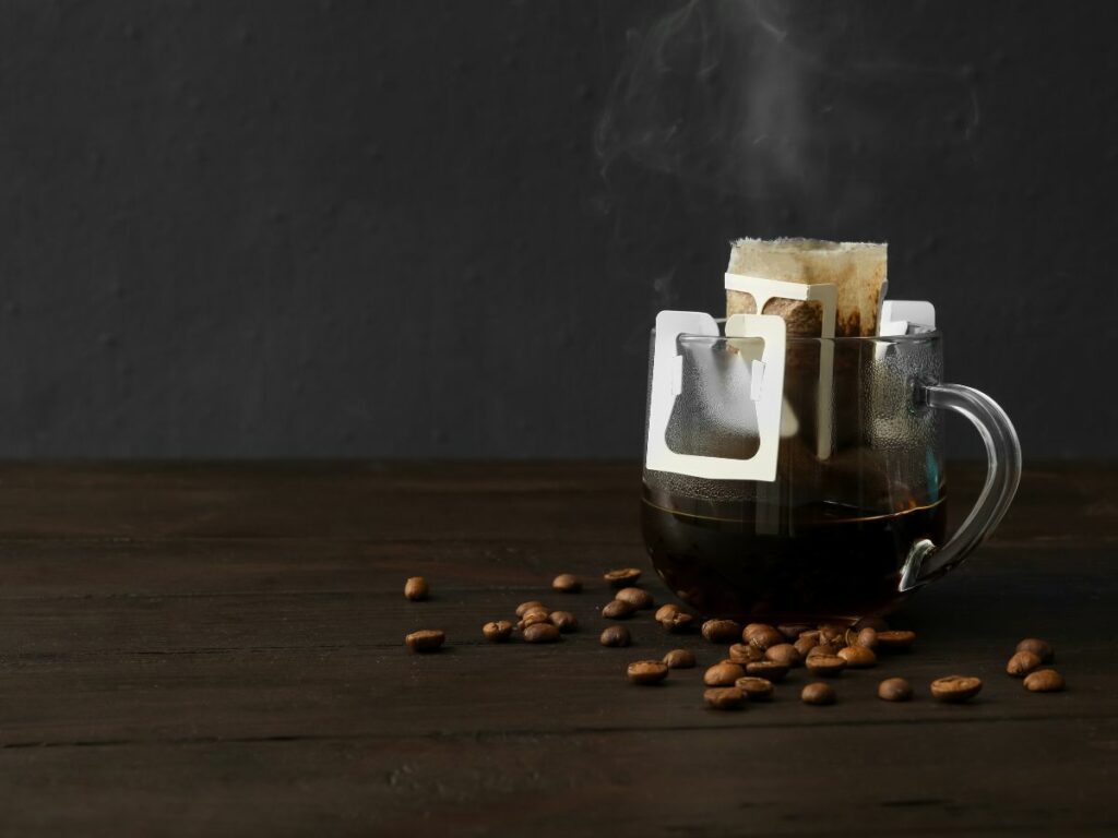 A cup of coffee with a tea bag filter and coffee beans around it.