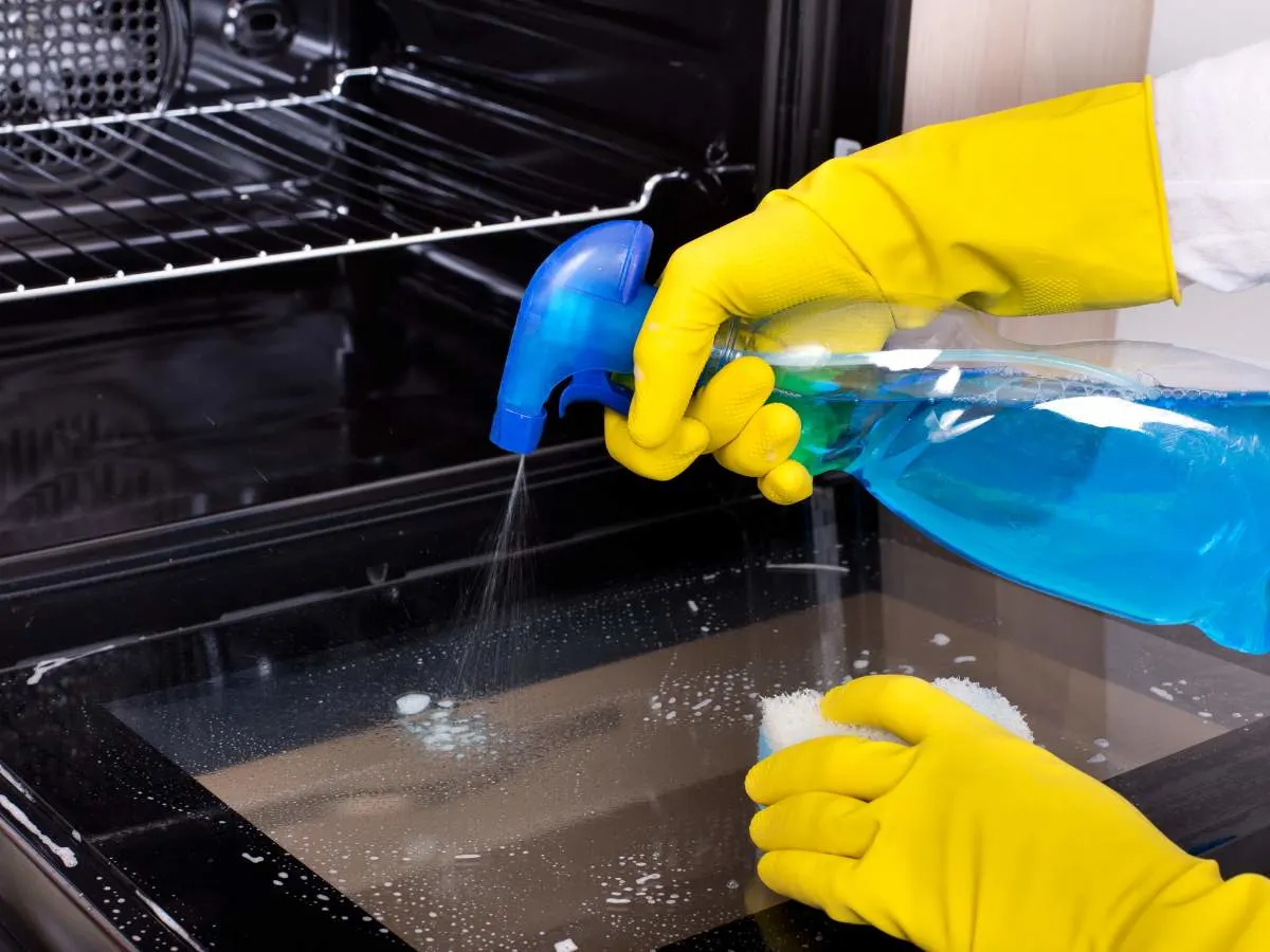 A person in yellow gloves cleaning an oven with a spray cleaner.