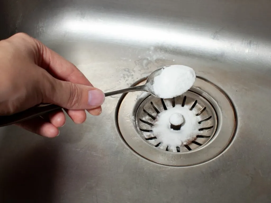 A person holding a spoon of baking soda over a sink drain.