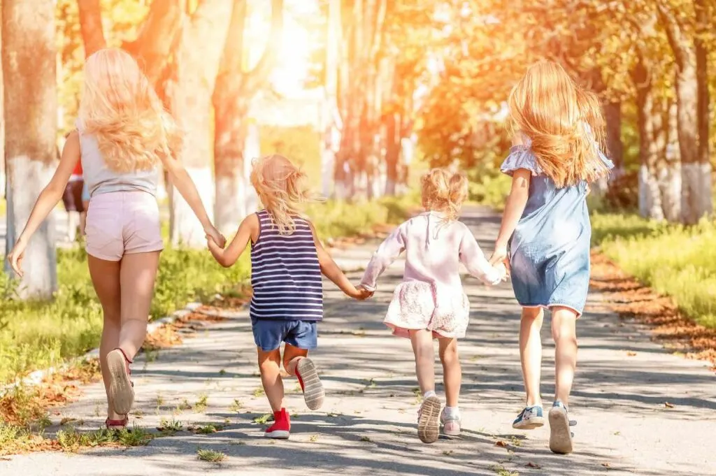 Four young girls holding hands walking down a road in a park.