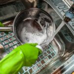 A person in green gloves washing a stainless steel pan in a kitchen sink.