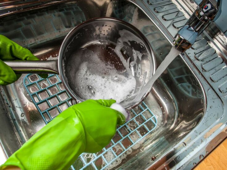 A person in green gloves washing a stainless steel pan in a kitchen sink.