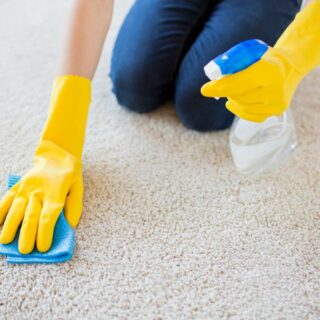 Person wearing yellow gloves cleaning a carpet with a spray bottle and cloth.