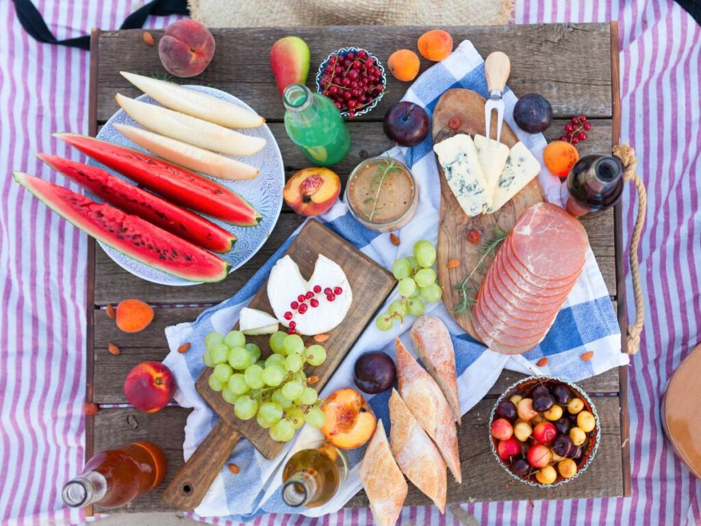 Picnic spread with an assortment of fruits, cheeses, bread, and cold cuts on a patterned blanket.