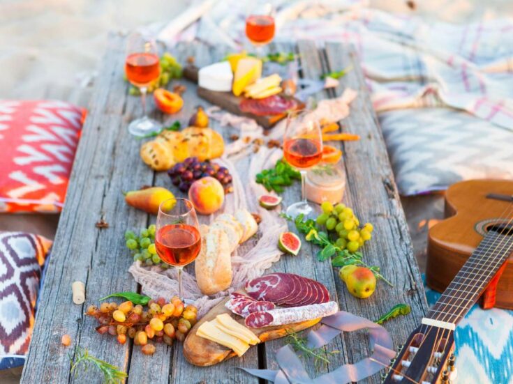 Beach picnic with a variety of snacks and wine on a charcuterie board, accompanied by a guitar.