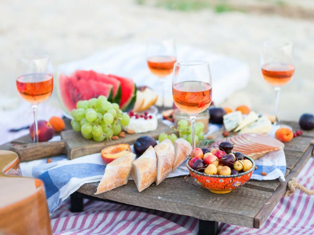 A summer charcuterie picnic setup with rosé wine, fresh fruit, and cheese on a wooden table at the beach.