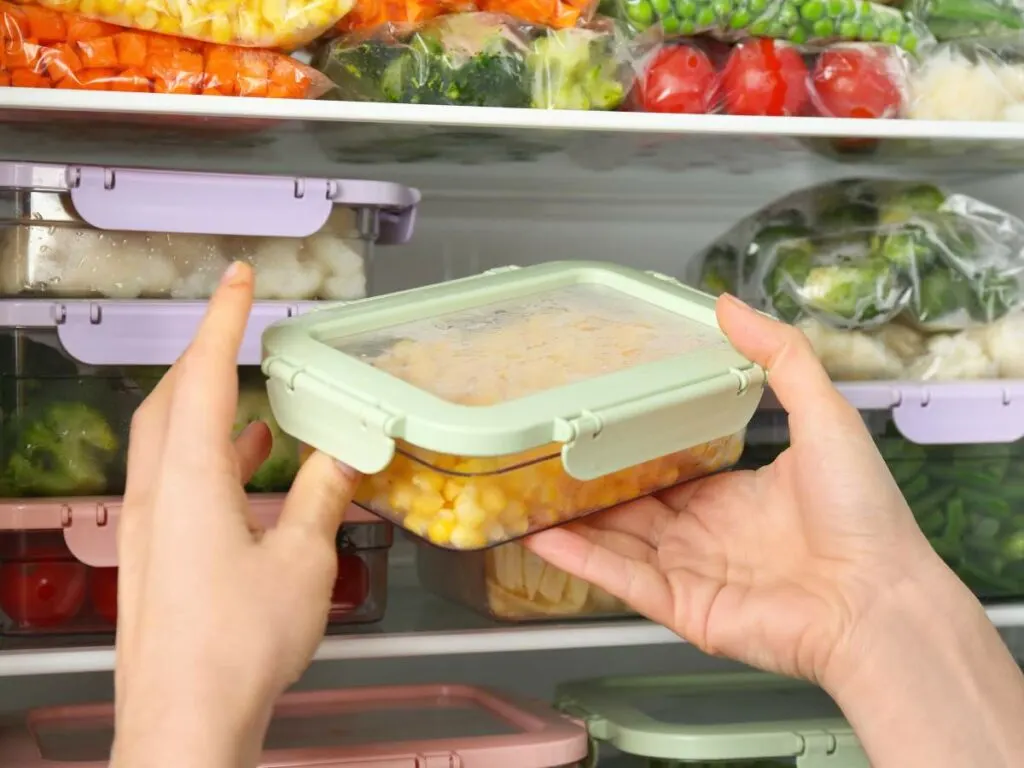 A person holding a plastic container full of vegetables in a refrigerator.