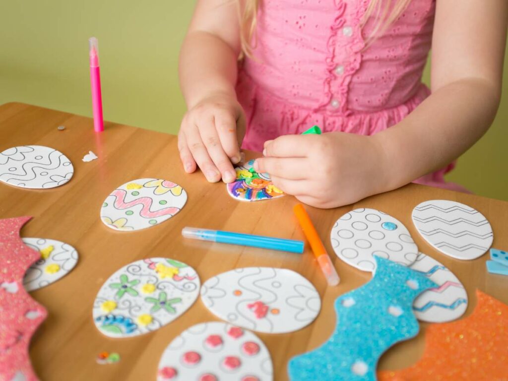 Child coloring easter egg cutouts on a table.