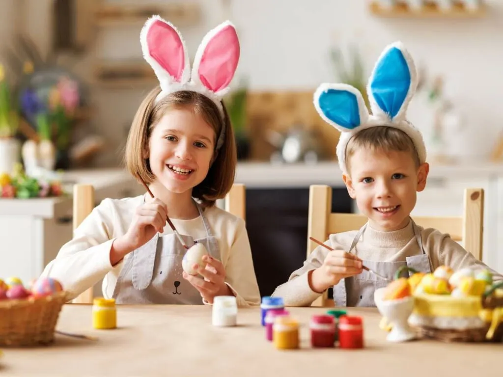 Two children wearing bunny ears painting easter eggs at a table.