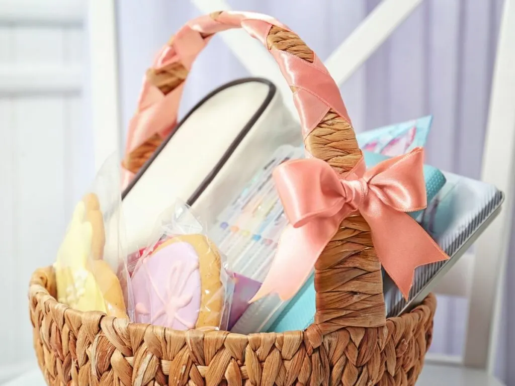 A wicker easter basket adorned with a pink ribbon, containing assorted items including cookies and a book.