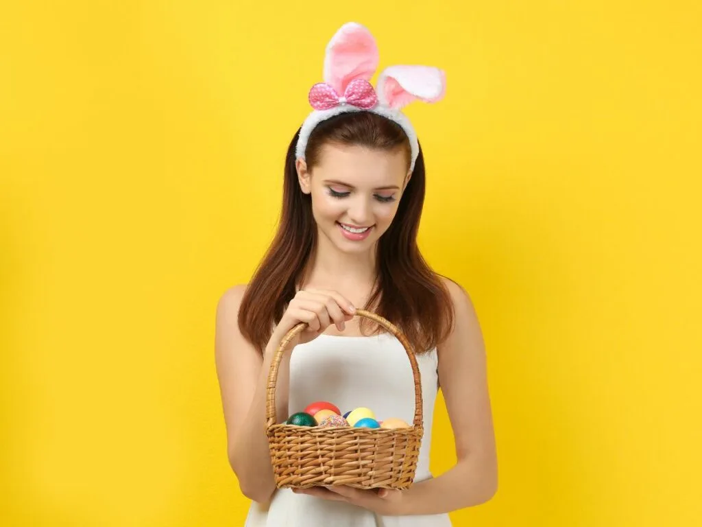 Young woman wearing bunny ears headband holding a basket of colorful easter eggs against a yellow background.