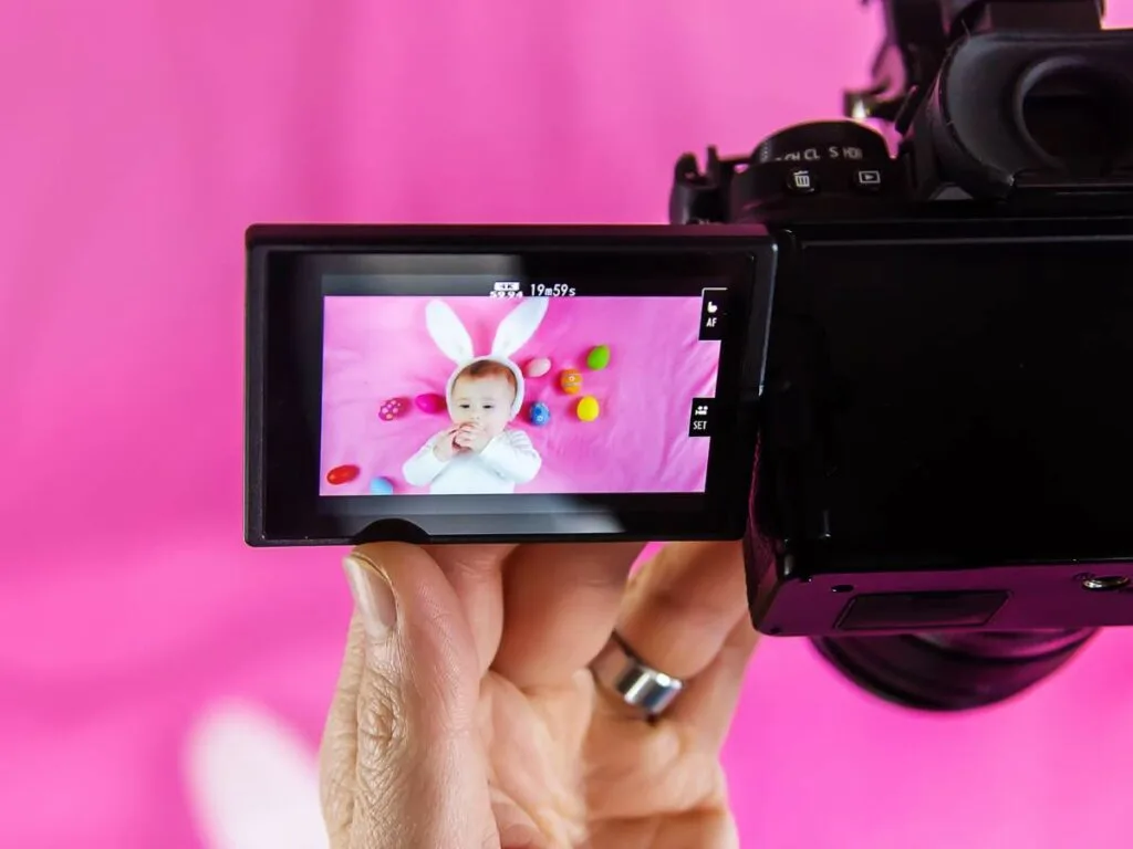 A person's hand holding a camera with a flip screen displaying a photo of a baby in a bunny costume against a pink background.