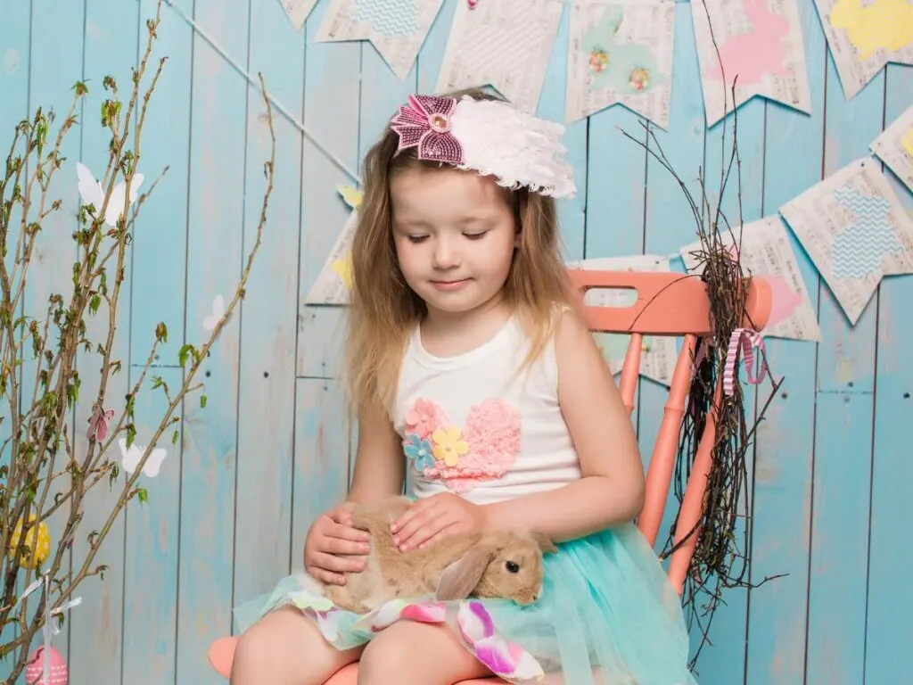 A young girl sitting on a chair, gently holding a rabbit, with a backdrop of pastel-colored decor and hanging paper Easter decorations.