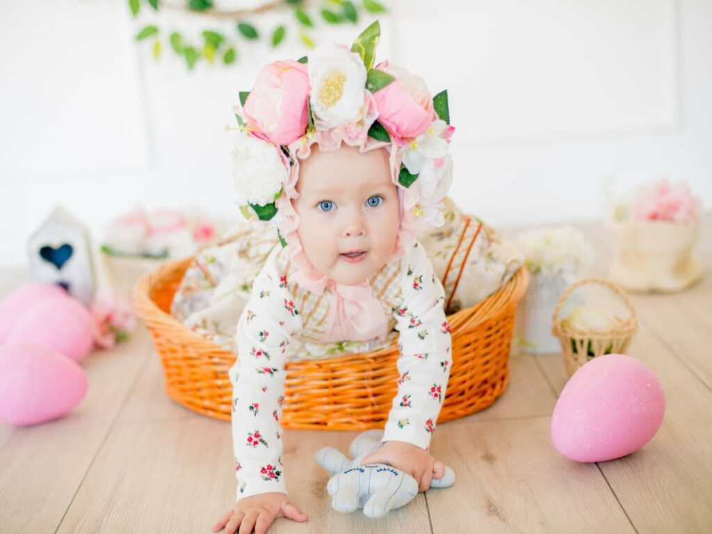 A baby with a floral headband crawls near a wicker basket and easter eggs.