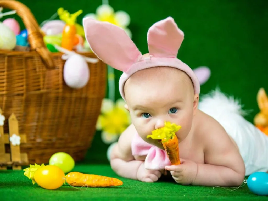 A baby wearing bunny ears and holding carrots with an Easter basket in the background.