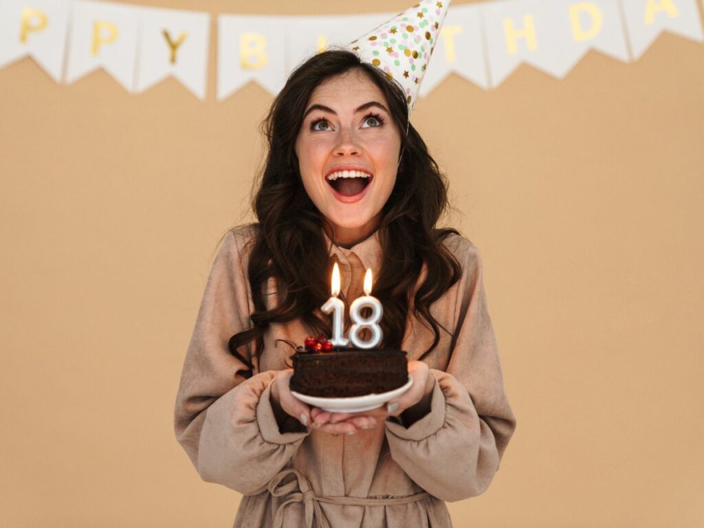 A woman in a party hat holding a birthday cake with lit candles that read "18", excitedly celebrating.