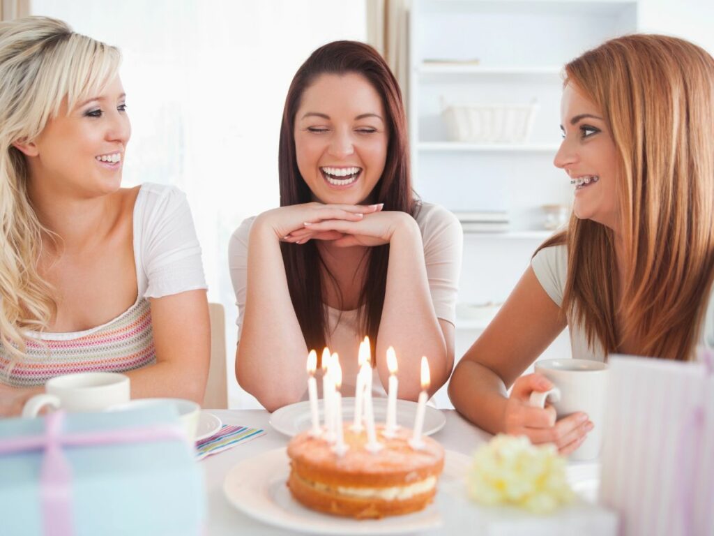 Three women laughing and enjoying a celebration with a small cake with lit candles on the table.