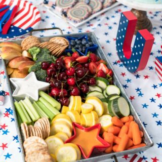A charcuterie board of assorted fruits and vegetables arranged with a patriotic theme on a star-spangled tablecloth.