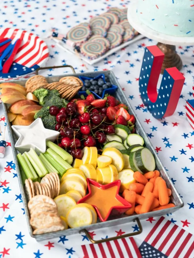 A charcuterie board of assorted fruits and vegetables arranged with a patriotic theme on a star-spangled tablecloth.