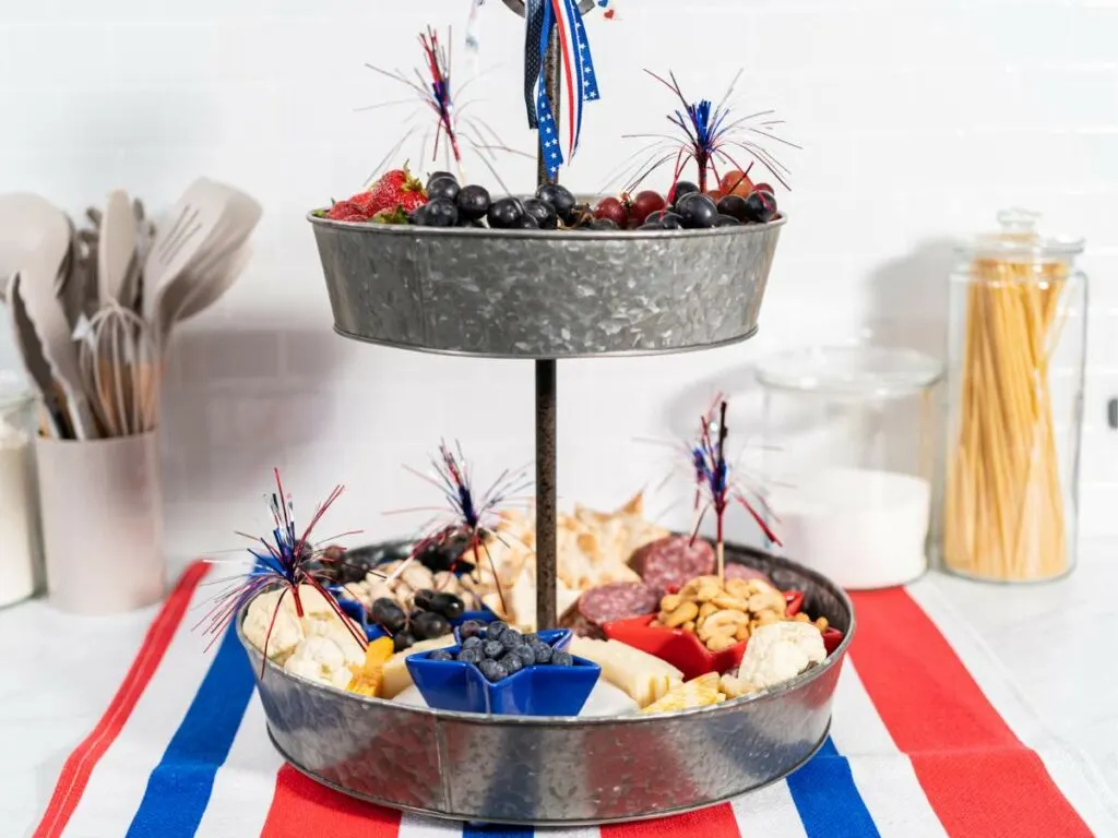 A two-tiered galvanized serving stand filled with a variety of fruits, cheese, and meats, decorated with patriotic ribbons and firework-style toppers, presented on a red, white, and blue striped cloth.