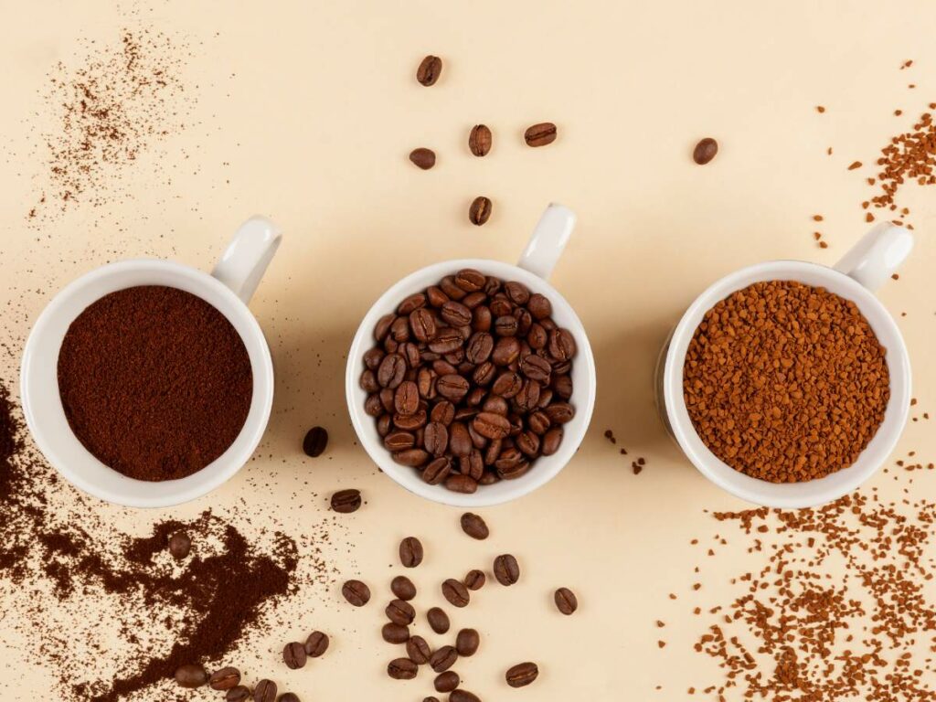 Three white cups containing different forms of coffee: ground coffee, whole beans, and instant coffee, arranged on a beige background with scattered coffee grounds and beans.