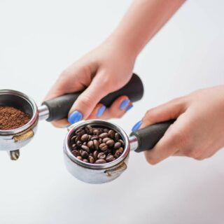 Two hands holding espresso portafilters filled with ground coffee and whole coffee beans.