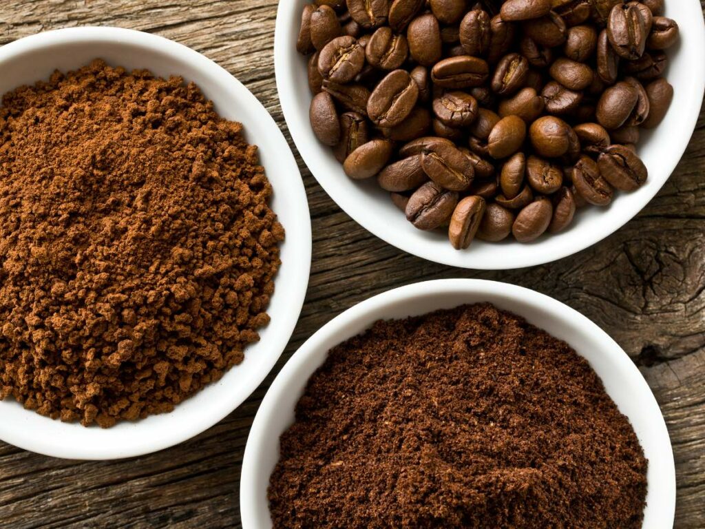 Three bowls containing different forms of coffee: ground, whole beans, and instant coffee, on a wooden surface.