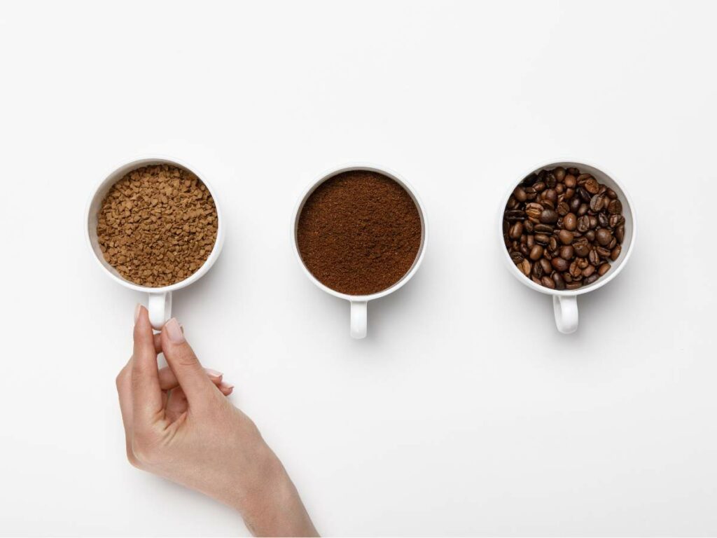 Three cups on a white surface showing different stages of coffee preparation: one with ground coffee, one with coffee powder, and one with coffee beans, with a hand touching the handle of the first cup.