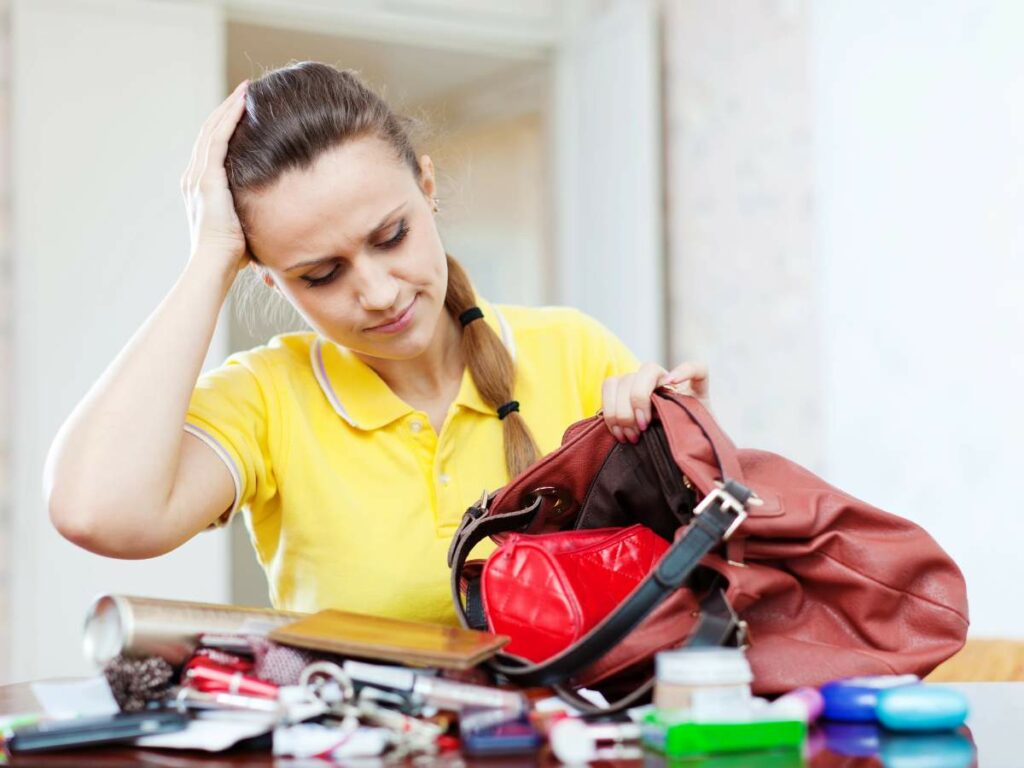 Woman in yellow shirt searching through a cluttered bag on a table filled with various items.